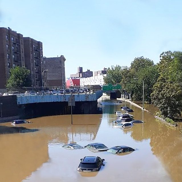 At left, tall apartment towers. At right, trees, In foreground, several mostly submerged cars in brown floodwaters, under a blue sky.
