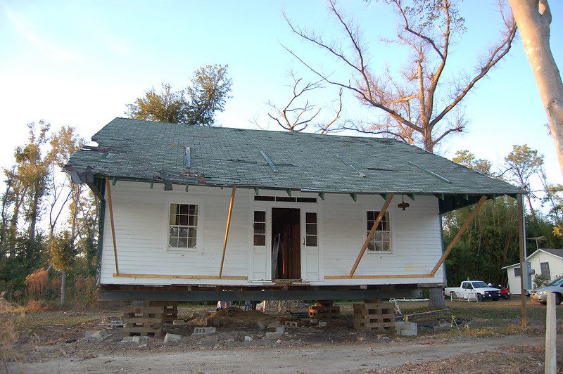 A small hurricane-damaged house is propped up on wooden crates. Long 2-by-4s hold up the roof over the front door. 