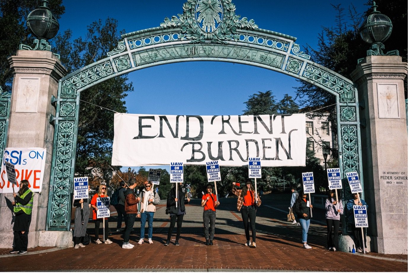 Under a blue sky, in a large arching gateway is suspended a banner that reads "End Rent Burden." Below it is a line of 11 picketers holding signs that say "UAW on Strike: Unfair Labor Practice." A passerby is talking to one of the picketers.