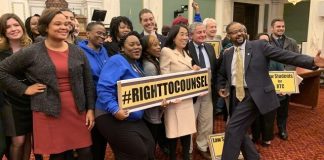 A group of about 30 people stand in a large room with marble architectural details. All are smiling broadly. Three are holding signs: one says "#RightToCounsel" and two others say "Law Students for RTC."