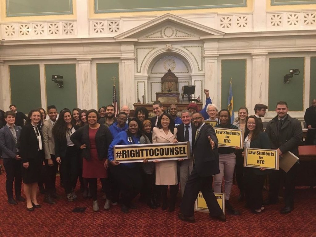 A group of about 30 people stand in a large room with marble architectural details. All are smiling broadly. Three are holding signs: one says "#RightToCounsel" and two others say "Law Students for RTC."