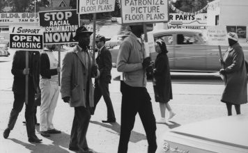 A black and white photo of seven people protesting racial discrimination in housing on a street corner, as a 1950s-era Buick drives past. The signs read "Stop racial discrimination now!"; "I support open housing"; "Don't patronize picture floor plans"; and a hand-lettered sign says "There can be no innocent bystanders." Most of the people in the photo are people of color; two are hidden by their signs.