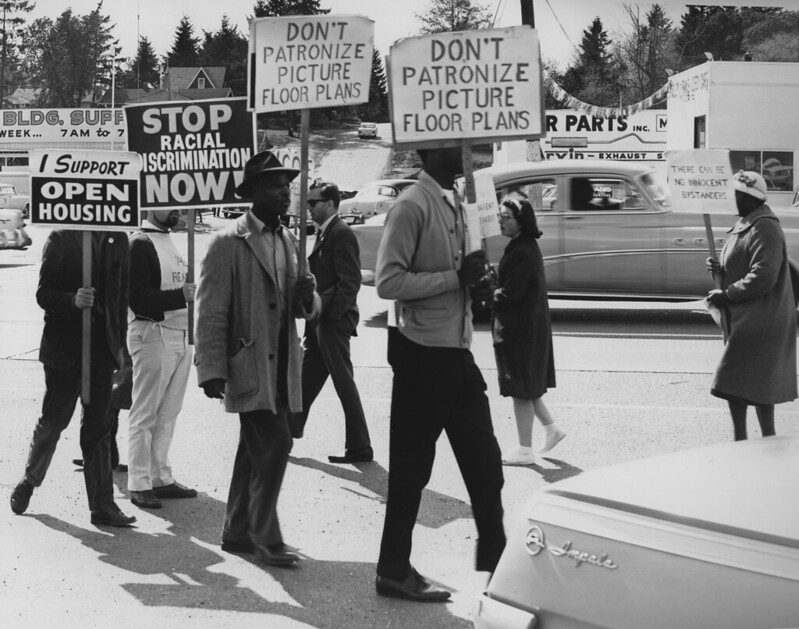 A black and white photo of seven people protesting racial discrimination in housing on a street corner, as a 1950s-era Buick drives past. The signs read "Stop racial discrimination now!"; "I support open housing"; "Don't patronize picture floor plans"; and a hand-lettered sign says "There can be no innocent bystanders." Most of the people in the photo are people of color; two are hidden by their signs.