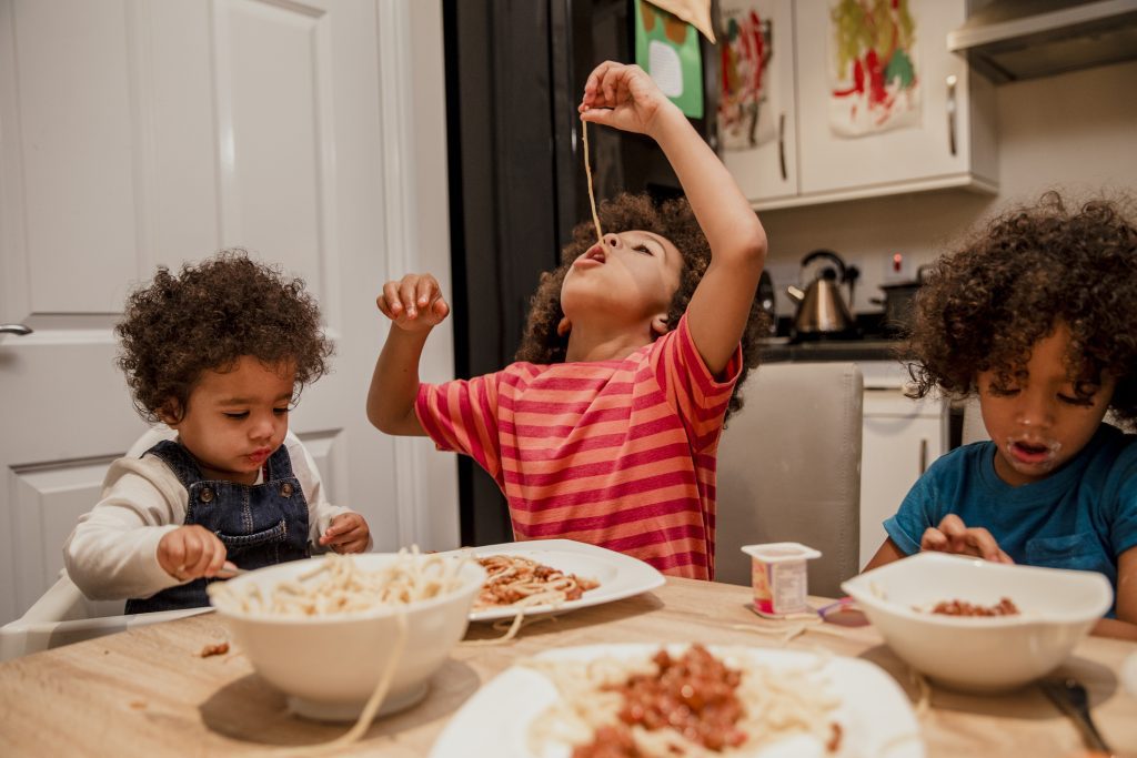 Three children are eating spaghetti at a kitchen table. One, wearing a striped shirt, is dangling a strand of spaghetti in the air over their mouth. The child on the right has a very messy face, and at left, a toddler in a high-chair is holding a spoon or fork and looking intently at their plate.