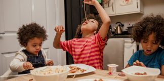 Three children eating spaghetti at a kitchen table. One, wearing a striped shirt, is dangling a strand of spaghetti in the air and eating it from the end. The child on the right has a very messy face, and at left, a child in a high-chair is holding a spoon or fork.