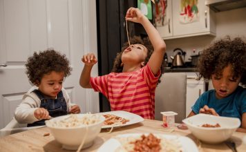 Three children eating spaghetti at a kitchen table. One, wearing a striped shirt, is dangling a strand of spaghetti in the air and eating it from the end. The child on the right has a very messy face, and at left, a child in a high-chair is holding a spoon or fork.