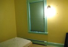 A room bare of furnishings except a mattress on the floor. The walls are yellow, the window frame is light green. The blinds are closed. There's a bare-bulb light fixture on the wall casting a glary light