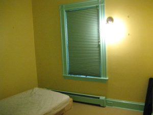 A room bare of furnishings except a mattress on the floor. The walls are yellow, the window frame is light green. The blinds are closed. There's a bare-bulb light fixture on the wall casting a glary light