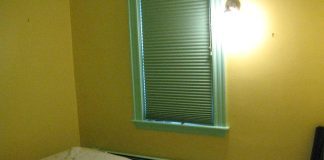 A room bare of furnishings except a mattress on the floor. The walls are yellow, the window frame is light green. The blinds are closed. There's a bare-bulb light fixture on the wall casting a glary light
