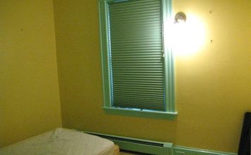 A room bare of furnishings except a mattress on the floor. The walls are yellow, the window frame is light green. The blinds are closed. There's a bare-bulb light fixture on the wall casting a glary light