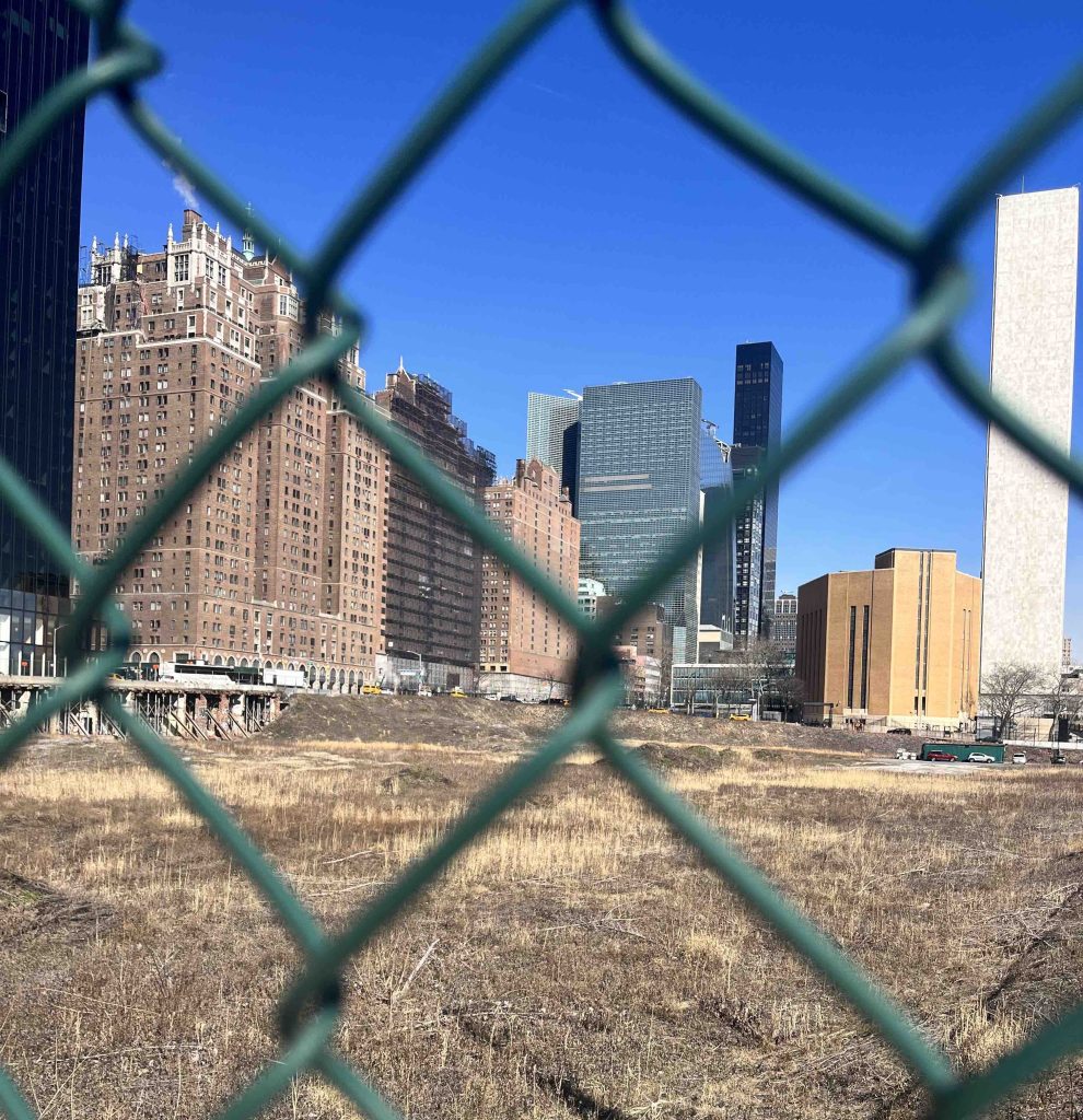 A view of an empty lot in Manhattan, seen through green chain-link fence. On the far side of the lot are tall brick buildings. At left is a tall black modern tower, and at right a tall white tower, all against a blue sky.