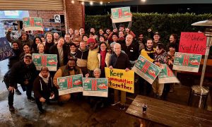 A large group of smiling people outdoors at night. Many are holding green and red rally signs in support of Measure H. Two handwritten signs read "Keep communities together" and "Stop unfair rent hikes."