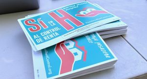 A pile of rally signs on a table. They read "Sí en la H al control de renta. The word "Sí" and the large letter H are both in red, other words are in white, all on a blue background. At right is a stylized image of red hand holding two white houses.