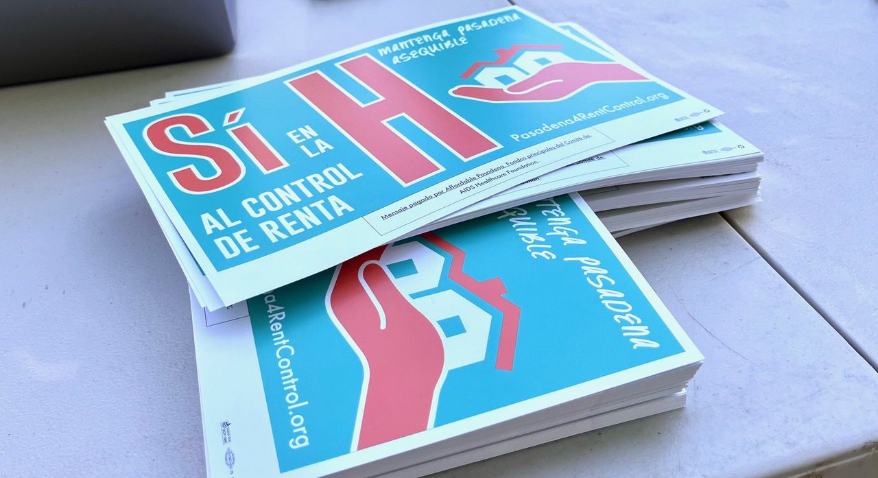 A pile of rally signs on a table. They read "Sí en la H al control de renta. The word "Sí" and the large letter H are both in red, other words are in white, all on a blue background. At right is a stylized image of red hand holding two white houses.