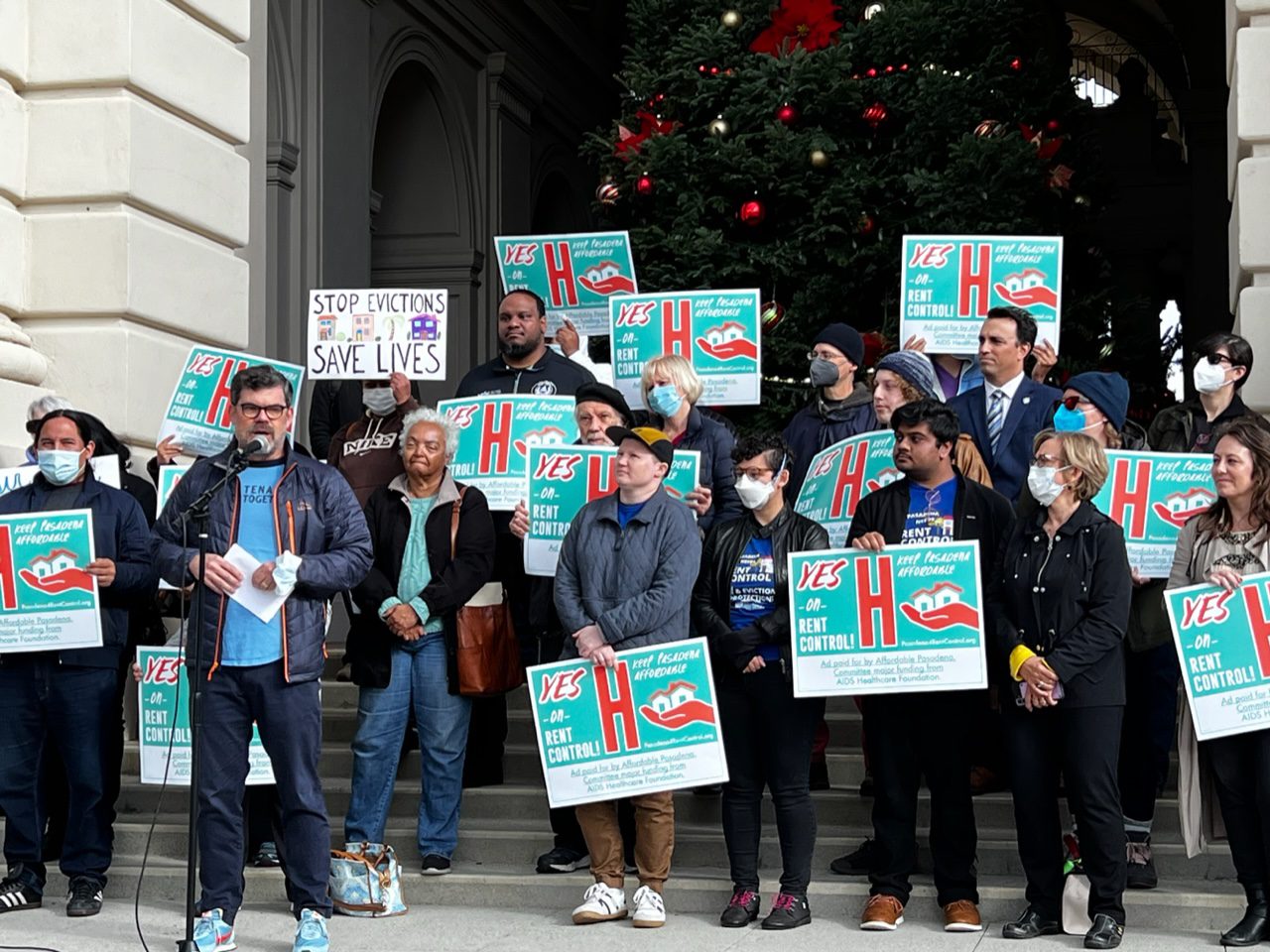 A group of people standing on steps, most holding signs saying "Yes on Rent Control" while at left a man speaks into a microphone. Behind the people, faintly visible, is a large holiday tree with red and silver ornaments.