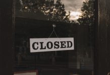 A sign saying "Closed" hands in a window that reflects treetops and the sky. The interior of the building is dark.