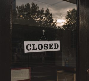 A sign saying "Closed" hands in a window that reflects treetops and the sky. The interior of the building is dark.