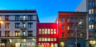 A two-story magenta building is sandwiched between two larger apartment buildings at dusk.