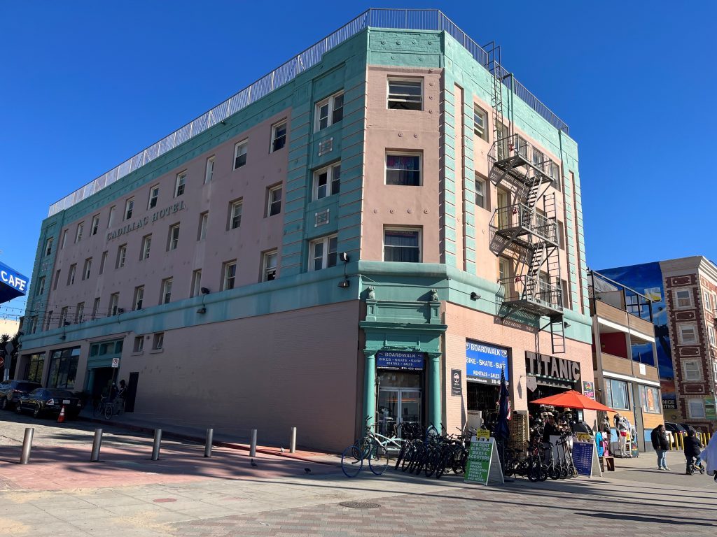 An early 1900s three- or four-story hotel on a street corner, seen from street level against a bright blue cloudless sky. Built of pink sandstone with light green trim. The ground floor has businesses; the sidewalk is crowded with parked scooters and a red cafe umbrella.