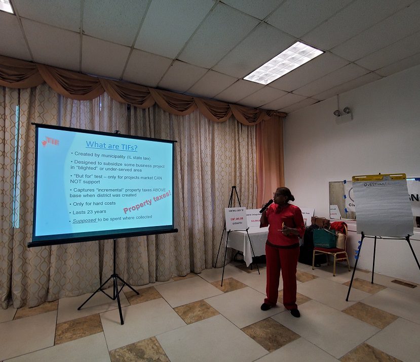 A dark-skinned woman in a red blouse stands holding a mic and looking at a screen on which is projected "What are TIFs?" with several bullet points beneath in much smaller type. Behind her is a curtain, and in the corner a table and chair with some meeting materials on them. The audience she is speaking to is not visible.