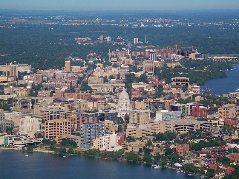 An aerial view of Madison, Wisconsin, with a lake in the foreground, the capital dome visible beyond it, and the city stretching beyond that.