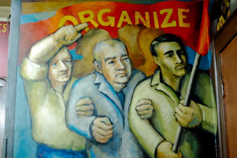 To illustrate the labor-housing coalition, a mural in reds, blues, pale greens, with black accents, show three men, arms linked, marching under a red banner that says "ORGANIZE" in yellow letters. The man on the left has his free arm upraised, finger pointing. 