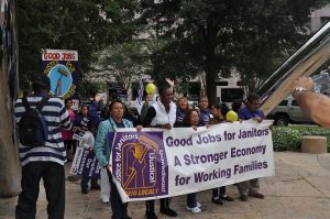 A large group of people march behind a large white banner with purple lettering that says "Good jobs for janitors: A stronger economy for working families"