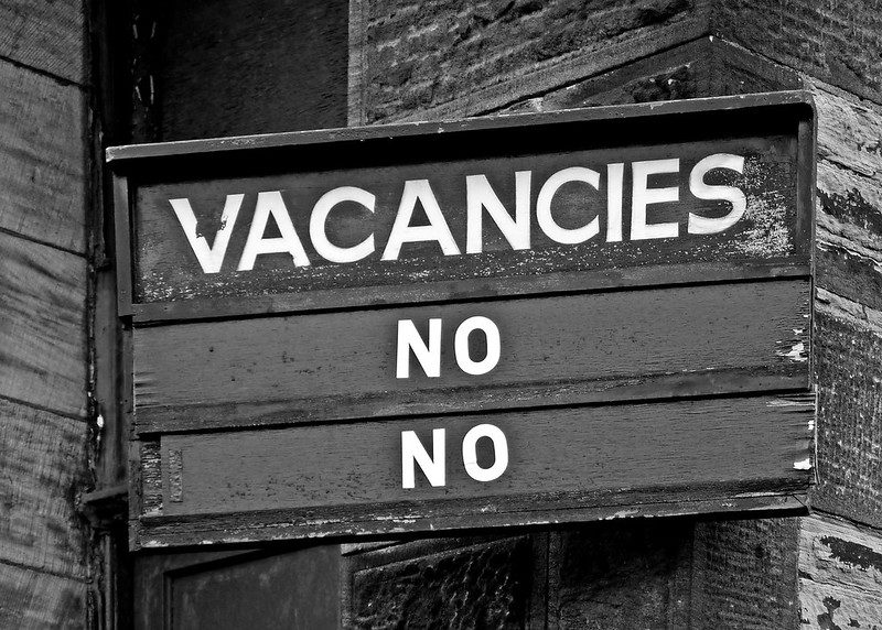 Closeup of a weatherbeaten wooden sign that says Vacancies in block letters across the top, and underneath that, No, and underneath that, another No.