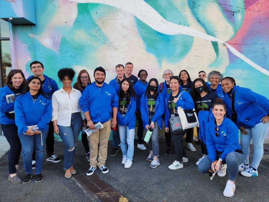 Nineteen smiling people of mixed ages and skin colors , most wearing blue sweatshirts, are standing (one is crouching) in front of a wall painted with an abstract mural in pastel colors.