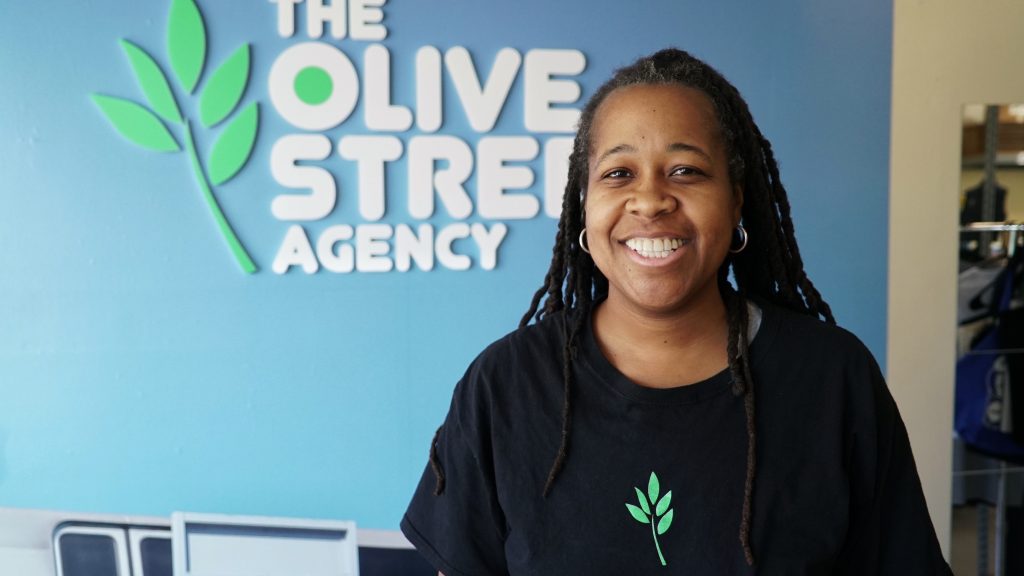 A broadly smiling woman with dark skin and braids, wearing a black shirt with a green-leaf logo on it, stands in front of a medium-blue wall. A business name, The Olive Street Agency, with the green-leaf logo, is printed (or affixed) to the wall in white capital letters.