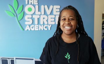A broadly smiling woman with dark skin and braids, wearing a black shirt with a green-leaf logo on it, stands in front of a medium-blue wall. A business name, The Olive Street Agency, with the green-leaf logo, is printed (or affixed) to the wall in white capital letters.