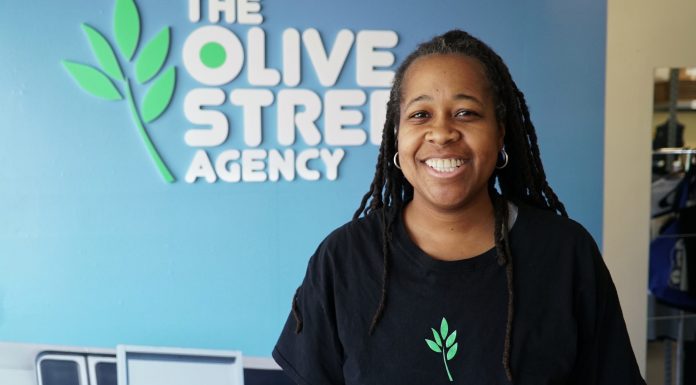 A broadly smiling woman with dark skin and braids, wearing a black shirt with a green-leaf logo on it, stands in front of a medium-blue wall. A business name, The Olive Street Agency, with the green-leaf logo, is printed (or affixed) to the wall in white capital letters.