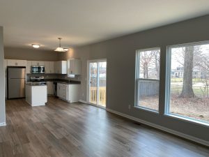 View of an open-plan first floor with a white-and-stainless steel kitchen at the far end. There's a sliding door to the back yard, and in the living area, a lare picture window with a view of a fenced yard.