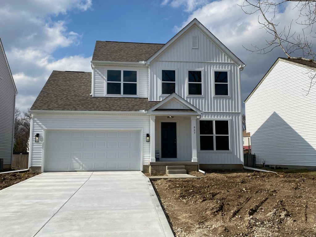 View from the end of the driveway of a new-looking white two-story house with a two-car garage, front porch with an overhang, and a gabled roof. The front yard is still all muddy soil with tire tracks.