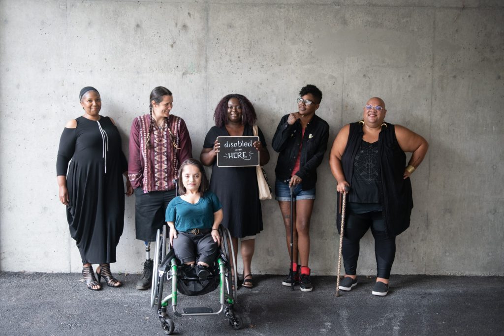 Six disabled people of color smile and pose in front of a concrete wall. Five people stand in the back, with the Black woman in the center holding up a chalkboard sign reading "disabled and here." A South Asian person in a wheelchair sits in front. Illustration for an article on laws regarding accessibility in housing.