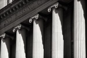 A tightly cropped black-and-white view of stone Ionic columns at the entrance to a courthouse. Words are carved on the lintel over the columns; visible in this photo are "and blessing."