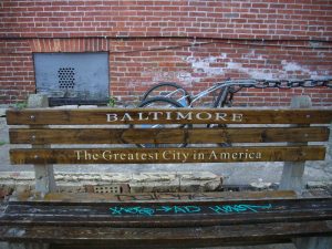 A park bench by a cracked sidewalk. There's graffiti on the bench seat, and the back is printed with "Baltimore/The Greatest City in America." Behind the bench is a brick wall with a gray metal vent at the left.