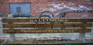 A park bench by a cracked sidewalk. There's graffiti on the bench seat, and the back is printed with "Baltimore/The Greatest City in America." Behind the bench is a brick wall with a gray metal vent at the left.