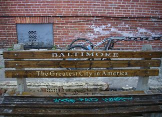 A park bench by a cracked sidewalk. There's graffiti on the bench seat, and the back is printed with "Baltimore/The Greatest City in America." Behind the bench is a brick wall with a gray metal vent at the left.