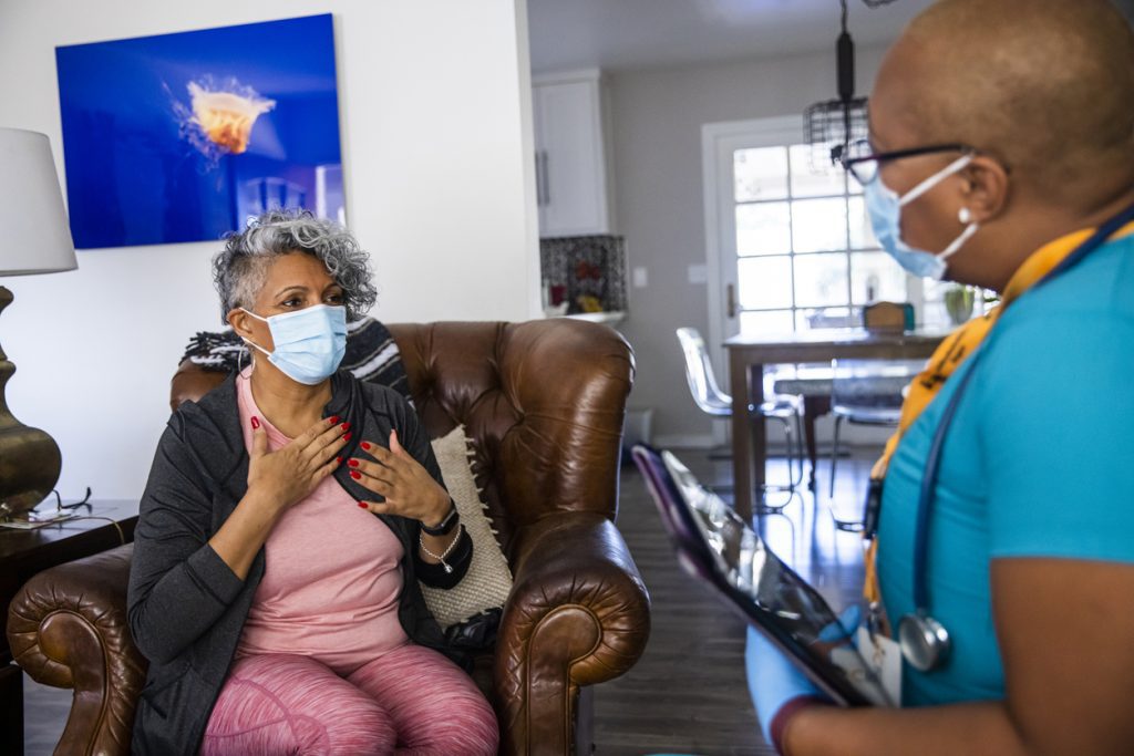 A senior Black woman wearing top and pants in shades of pink with a dark gray sweater, and wearing a surgical mask, sits in an armchair facing a home healthcare nurse. She is Black, with very short hair and wearing black-rimmed glasses, blue scrub top; also wearing a surgical mask and stethoscope, and carrying a clipboard or tablet. On the wall behind the seated woman is a bright blue artwork.