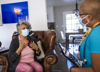 A senior Black woman wearing top and pants in shades of pink with a dark gray sweater, and wearing a surgical mask, sits in an armchair facing a home healthcare nurse. She is Black, with very short hair and wearing black-rimmed glasses, blue scrub top; also wearing a surgical mask and stethoscope, and carrying a clipboard or tablet. On the wall behind the seated woman is a bright blue artwork.