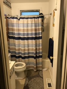 A bathroom seen before modifications were done to make it accessible. A shower curtain in stripes of gray, black, and tan partially conceals a standard bathtub, with its high wall presenting a barrier.