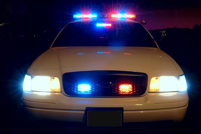 A close nighttime view of the front of a squad car, with red and blue lights glowing on the roof, and red and blue lights in the grille. The headlights are also on. The windshield appears black and no people are visible in the car.