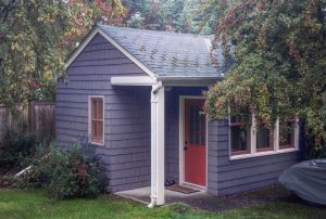 A small, one-story, gray-shingled cottage with a red door and white trim, with overhanging trees on the left and right.