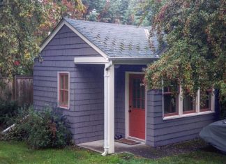 A small, one-story, gray-shingled cottage with a red door and white trim, with overhanging trees on the left and right.