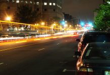 A nighttime photo in Jersey City, NJ. The photo is take on the side of a street, with orange light trails in the center of the image going down the street. There are parked cars on the right side of the image, and high-rise buildings to the left.