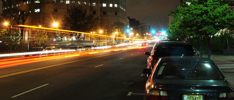 A nighttime photo in Jersey City, NJ. The photo is take on the side of a street, with orange light trails in the center of the image going down the street. There are parked cars on the right side of the image, and high-rise buildings to the left.