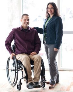 In a formal posed photo, a young middle-aged white man sits in a wheelchair with his right hand on the wheel. He has short hair and is wearing a maroon shirt and khaki pants. Standing next to him, with her hand on his shoulder, is a similar-aged woman with medium length brown hair and wearing a light green shirt, dark green sweater, and gray slacks. They're both smiling. Behind them is a blurry and indistinct view through a floor-to-ceiling window.