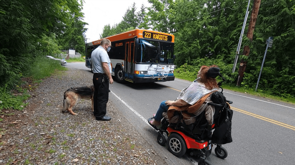 A woman in a motorized wheelchair travels along a rural road that has no sidewalks, as a bus approaches close to her from the opposite direction. Standing on the coarse gravel shoulder of the road is a man with a dog on a short leash. 