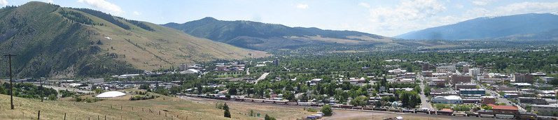 A distant panoramic view of the city of Missoula, Montana. The city is in the right foreground and at left, and beyond the city, rise mountains, under a pale blue sky with occasional clouds.
